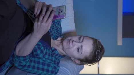 Vertical-video-of-Woman-lying-on-the-sofa-at-night-texting-on-the-phone.-Happy-and-in-good-spirits.